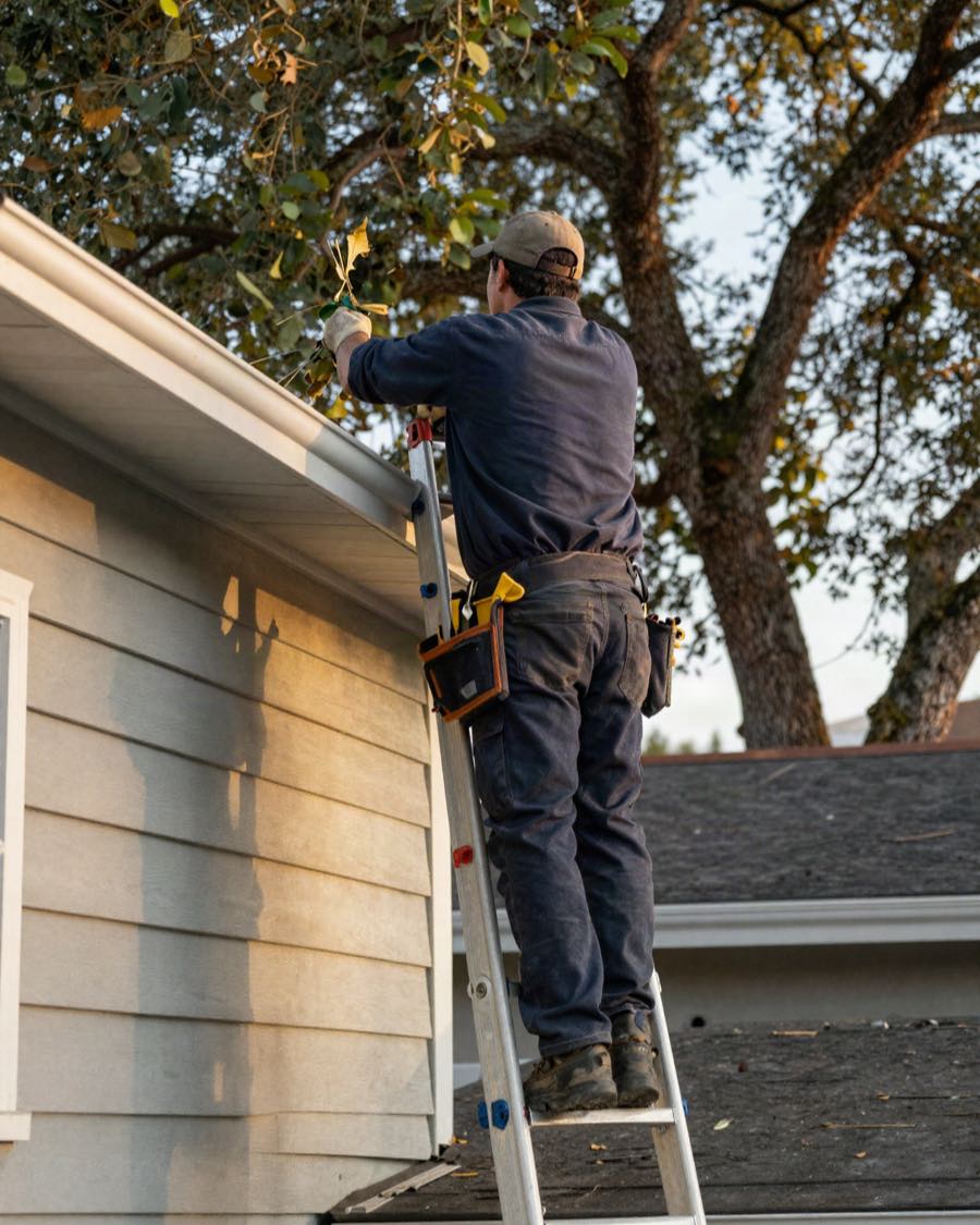 A contractor on a ladder at the roofline of a house, pulling leaf debris out of the gutter by hand.